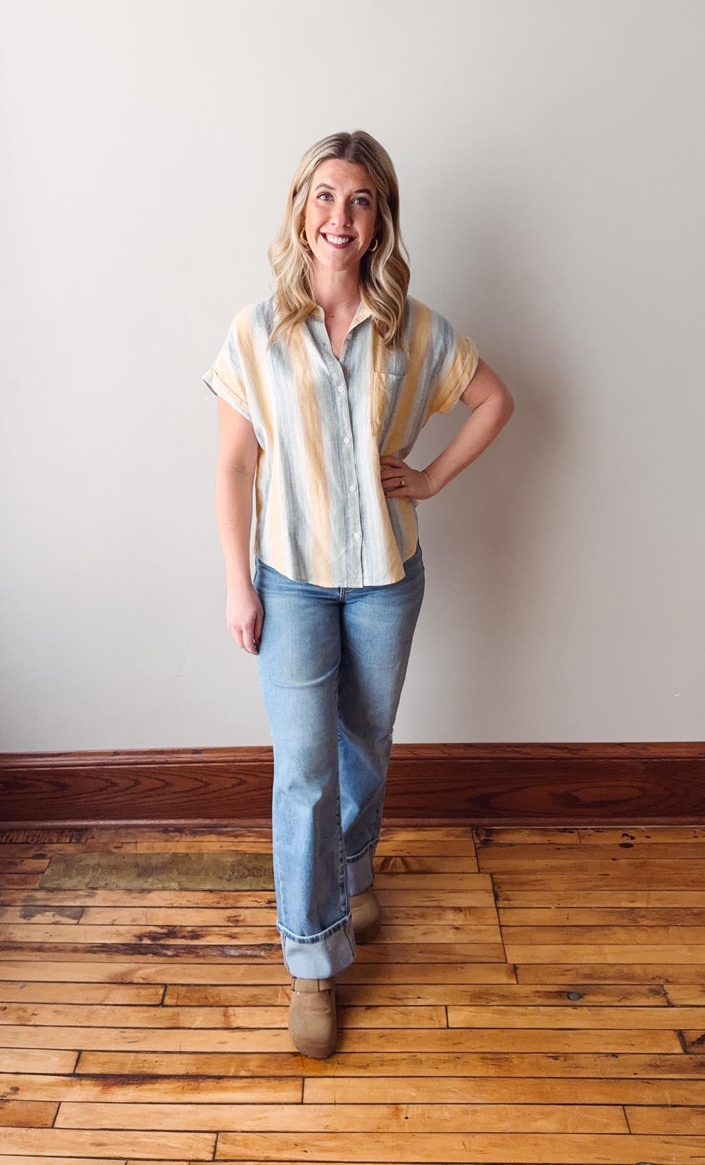 Woman wearing a striped shirt and jeans standing on a wooden floor with a white wall background
