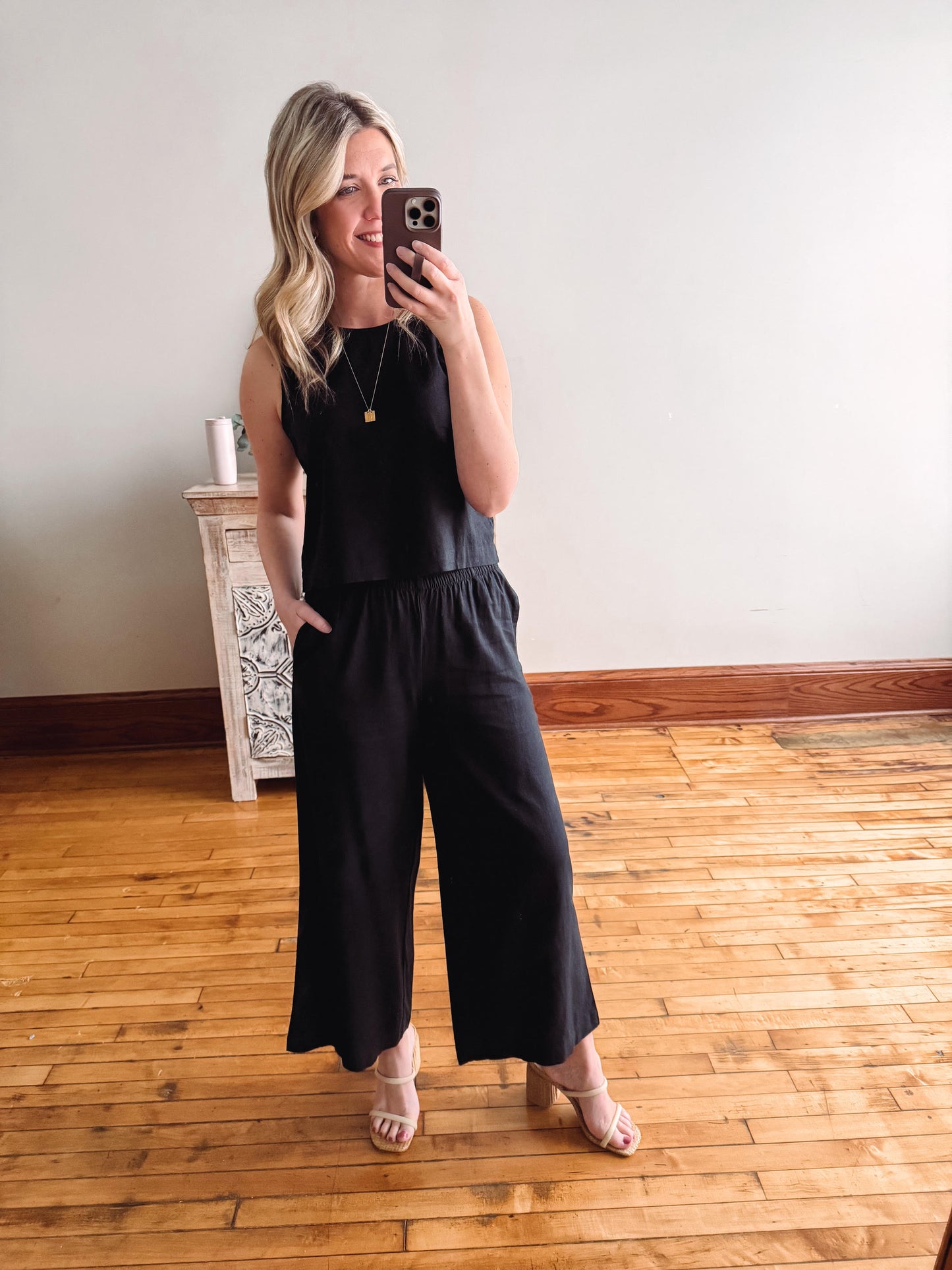 Woman taking a mirror selfie wearing a black jumpsuit in a room with wooden flooring.
