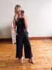 Woman taking a mirror selfie wearing a black jumpsuit in a room with wooden flooring.