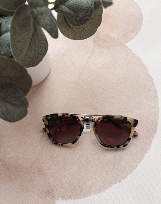 Sunglasses with tortoiseshell frame and dark lenses on a pink surface with green leaves.