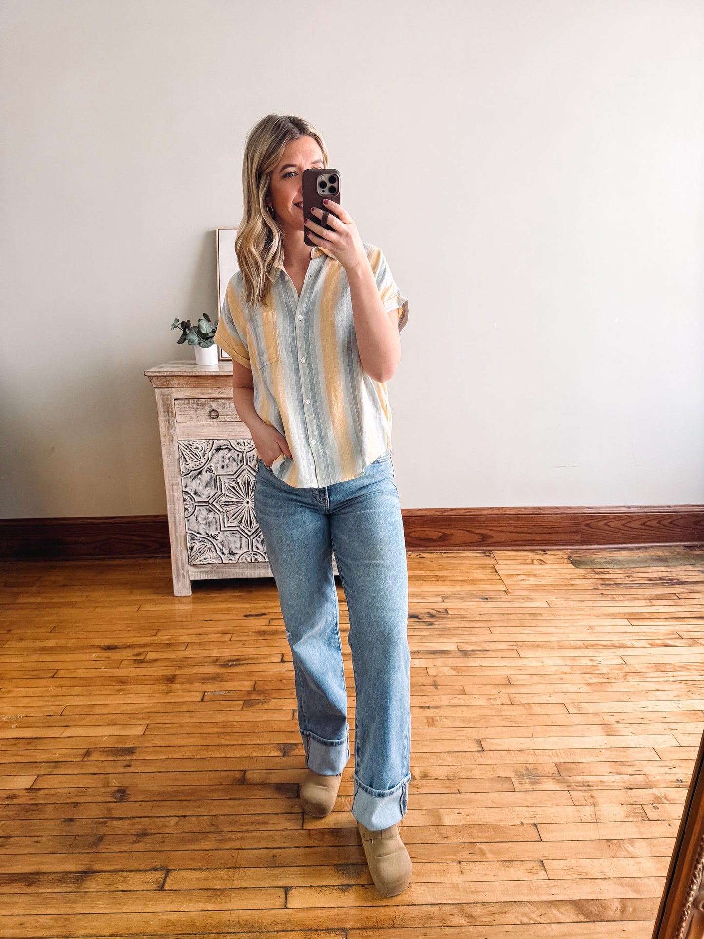 Person taking a mirror selfie wearing a striped shirt and jeans in a room with wooden flooring.
