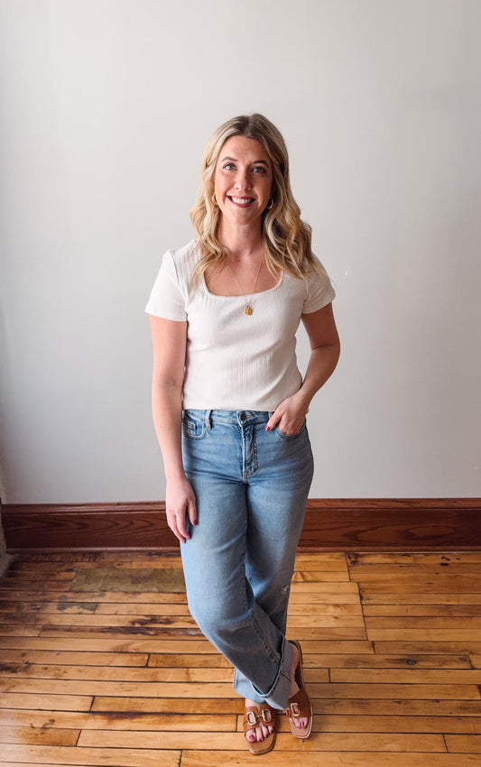Woman wearing a white top and blue jeans standing on a wooden floor with a plain wall background