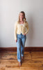 Woman wearing a yellow cardigan and blue jeans standing on a wooden floor against a white wall.