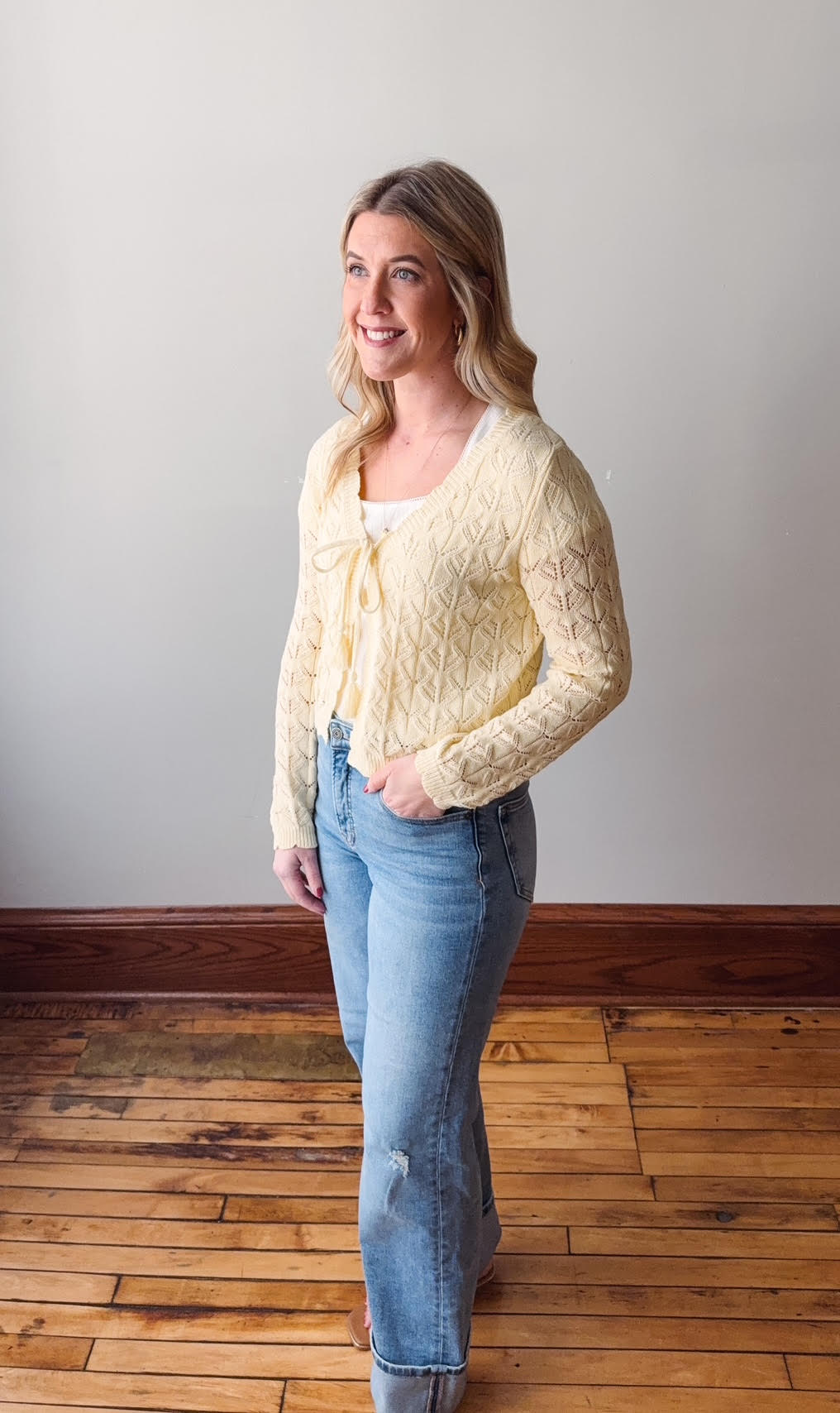 Woman wearing a yellow lace cardigan and blue jeans standing on a wooden floor.