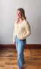 Woman wearing a yellow lace cardigan and blue jeans standing on a wooden floor.