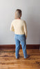 Person wearing a yellow knitted sweater and blue jeans standing on a wooden floor against a white wall.