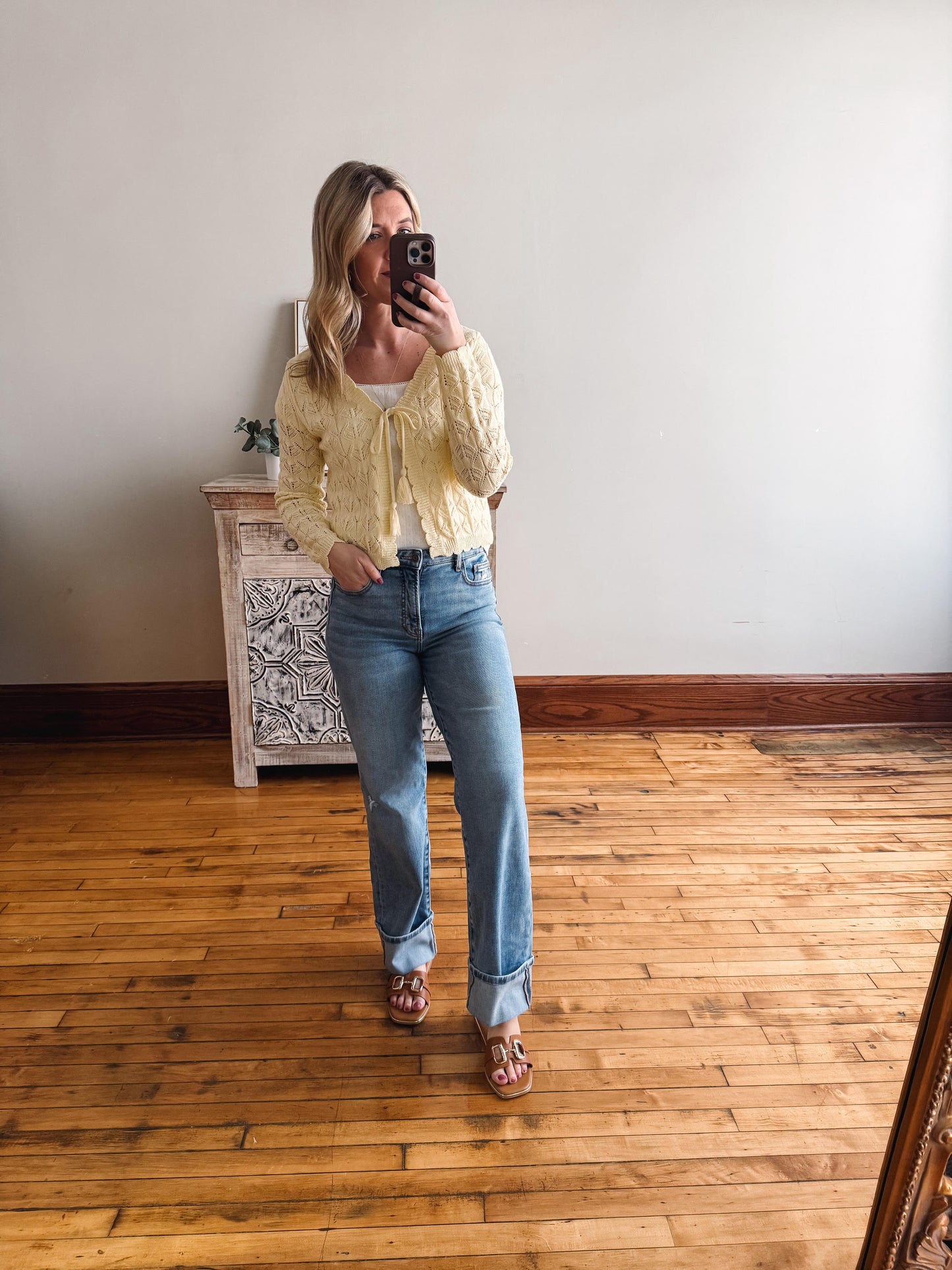 Woman taking a mirror selfie wearing a yellow cardigan and blue jeans in a room with wooden flooring.