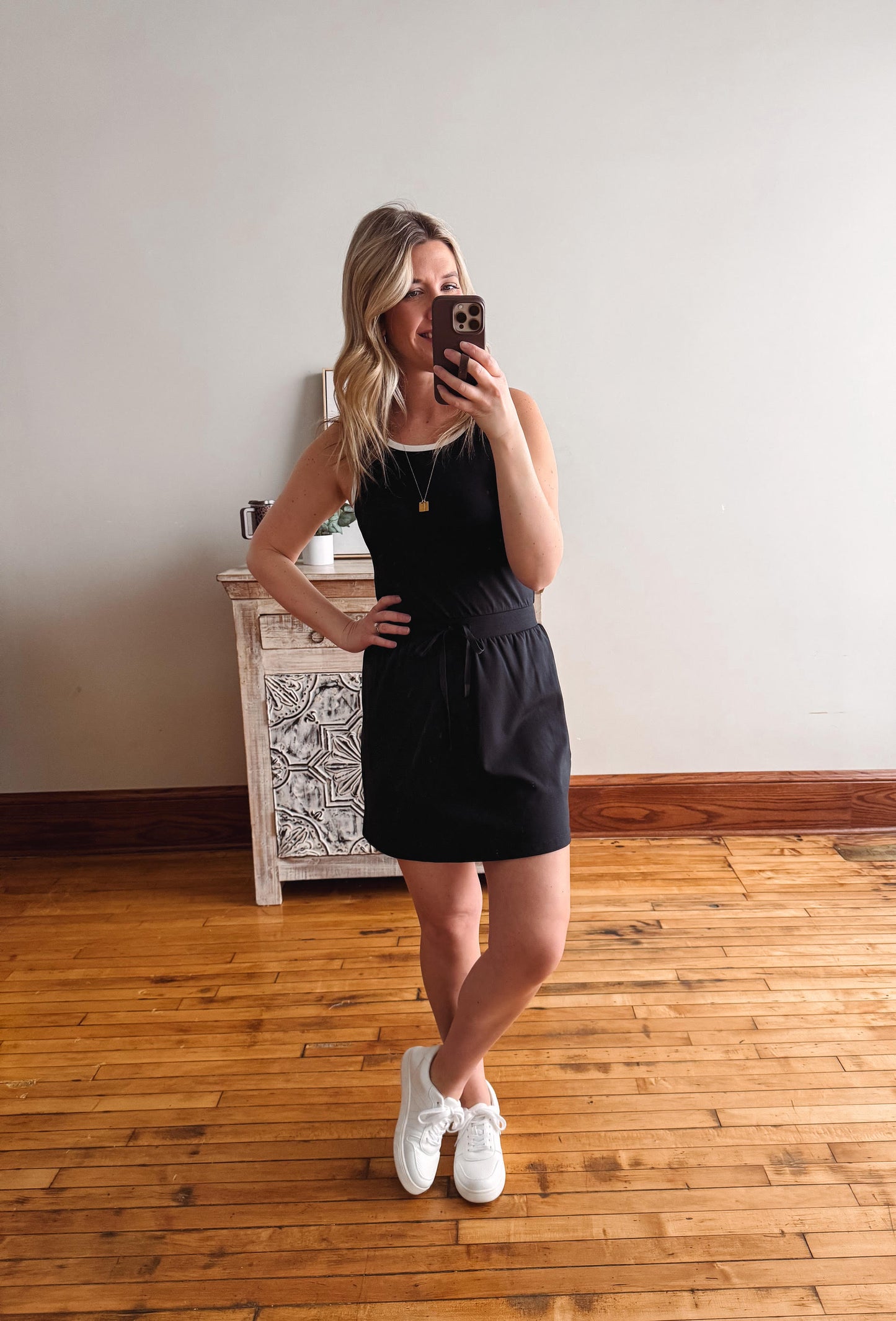 Woman taking a mirror selfie wearing a black dress and white sneakers in a room with wooden flooring.